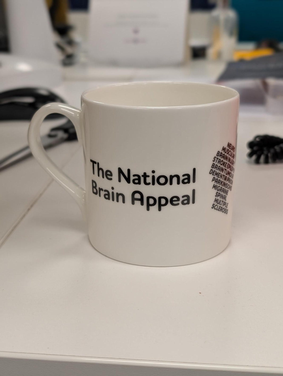 White ceramic mug on a desk showing The National Brain Appeal logo and a list of neurological conditions printed on the side.