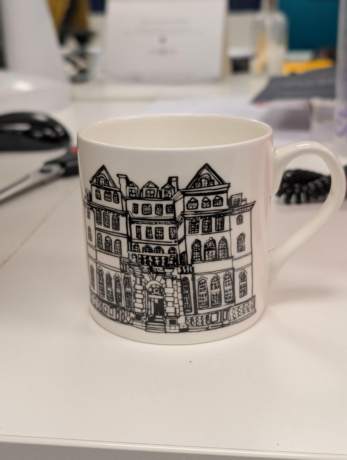 White ceramic mug on a desk, printed with a black line illustration of a historic building façade.